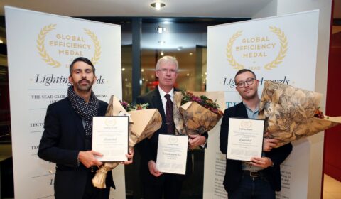 Three men in suits hold bouquets and certificates in front of Global Efficiency Medal banners, posing for a photo at an award ceremony.