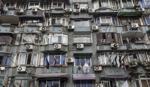 Close-up of an apartment building with many windows, air conditioning units, laundry hanging out to dry, and some balconies enclosed or covered with awnings.