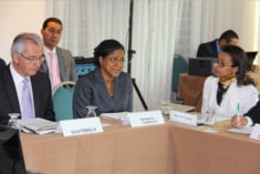Four people in formal attire sit at a conference table with nameplates and documents, engaged in discussion. Two men are in the background, and three people are facing the camera in the foreground.
