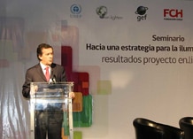 A man in a suit speaks at a clear podium during a seminar. Banners and logos are visible on the wall behind him with Spanish text about lighting strategies and project results.