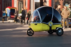 A small, yellow three-wheeled vehicle with a bubble-shaped transparent canopy drives on a city street while people walk in the background.