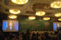 A conference room with chandeliers, a panel of speakers on stage, and a large screen displaying a person speaking, at the Global Efficient Lighting Forum.