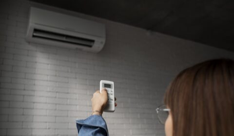 A person holding a remote control and pointing it at a wall-mounted air conditioner unit in a room with white brick walls.