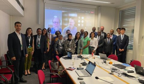 A group of professionals pose for a photo in a conference room, with laptops on the table and others joining via video call on a large screen in the background.