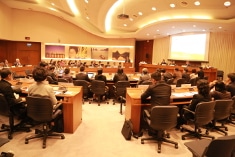 People in formal attire sit around a large conference room table, listening to a presentation. There are images on the wall, and a projection screen is lit at the front of the room.