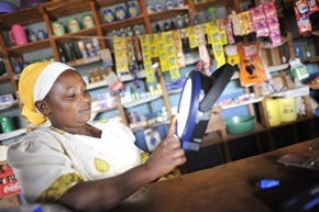 A woman in a yellow headscarf uses a handheld device while sitting behind a counter in a small shop filled with colorful products on shelves.