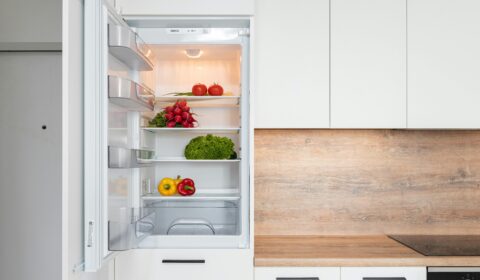 Open refrigerator in a modern kitchen, containing radishes, lettuce, and bell peppers on the shelves. The surrounding cabinets are white, and the countertop is wood-colored.