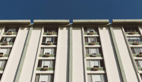 A tall apartment building with vertical lines, balconies, and window air conditioning units, photographed from below against a clear blue sky.