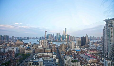 Cityscape view of Shanghai with the Huangpu River, tall modern skyscrapers including the Oriental Pearl Tower, and densely packed older buildings in the foreground under a blue sky.