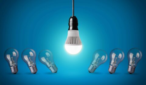 A glowing LED bulb hangs above five unlit traditional lightbulbs, which are arranged in a semicircle on a blue background.