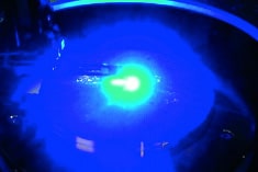 A bright blue laser beam illuminates a dark surface, creating a glowing circular pattern with a bright center and scattered light reflections.