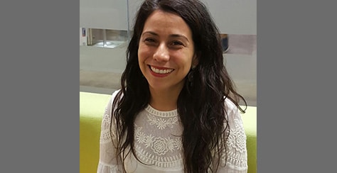 A woman with long dark hair, wearing a white lace top, smiles while sitting on a light green couch. The background is blurred, with glass panels and a hallway visible.