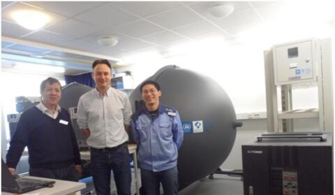 Three men stand in a laboratory with a large cylindrical machine and electronic equipment behind them. The setting appears to be scientific or industrial, with bright overhead lighting.