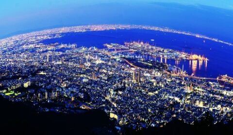 A vibrant cityscape at dusk with numerous lights, tall buildings, and a large body of water in the background, viewed from a high vantage point.