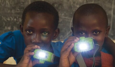Two children in blue shirts hold bright headlamps, shining them toward the camera, with a chalkboard in the background.