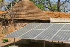 A solar panel array stands outdoors in front of a thatched-roof building with trees and stone walls in the background.