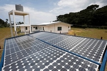 Rows of solar panels are installed outdoors in front of a white building with a water tank tower, surrounded by grass and trees under a partly cloudy sky.