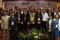 A large group of professionally dressed people pose together indoors at a conference with a banner behind them displaying various organizational logos.