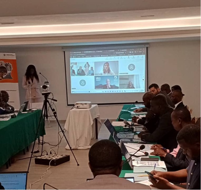 A woman presents at a conference room with participants seated at tables using laptops, while a virtual meeting with several people is projected on a screen behind her.