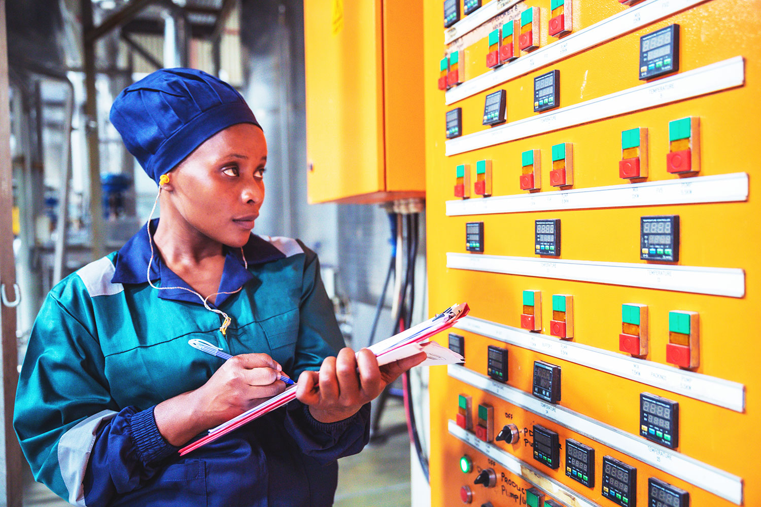 A woman in workwear and a blue headscarf holds a clipboard and pen, inspecting and taking notes on a large control panel with many buttons and digital displays in an industrial setting.