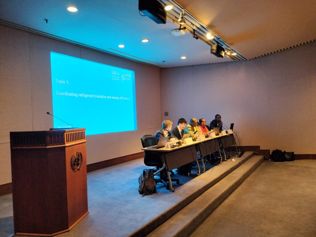 Five people sit at a long table with laptops during a conference in a room with a blue presentation slide reading Coordinating refrigerant transition and energy efficiency. A wooden podium is on the left.