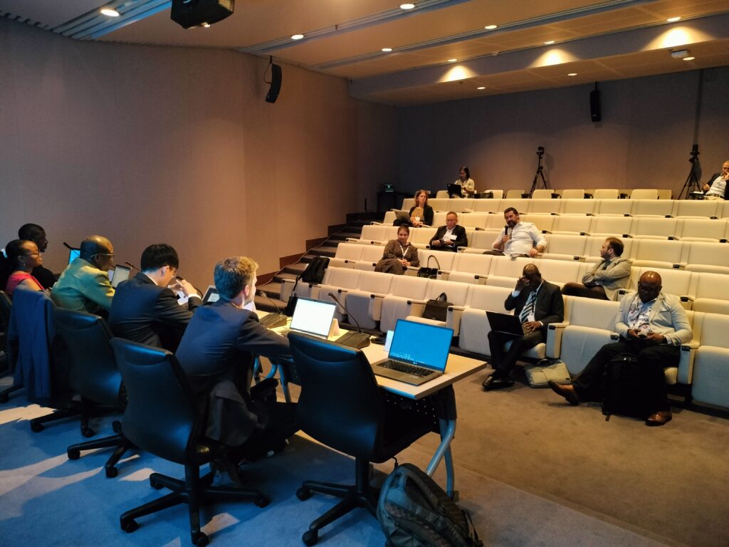 A small group of people sits spaced out in a conference room with tiered seating. Several attendees use laptops while others listen to speakers at a panel table in front.