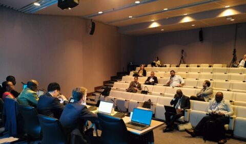 A small group of people sits spaced out in a conference room with tiered seating. Several attendees use laptops while others listen to speakers at a panel table in front.